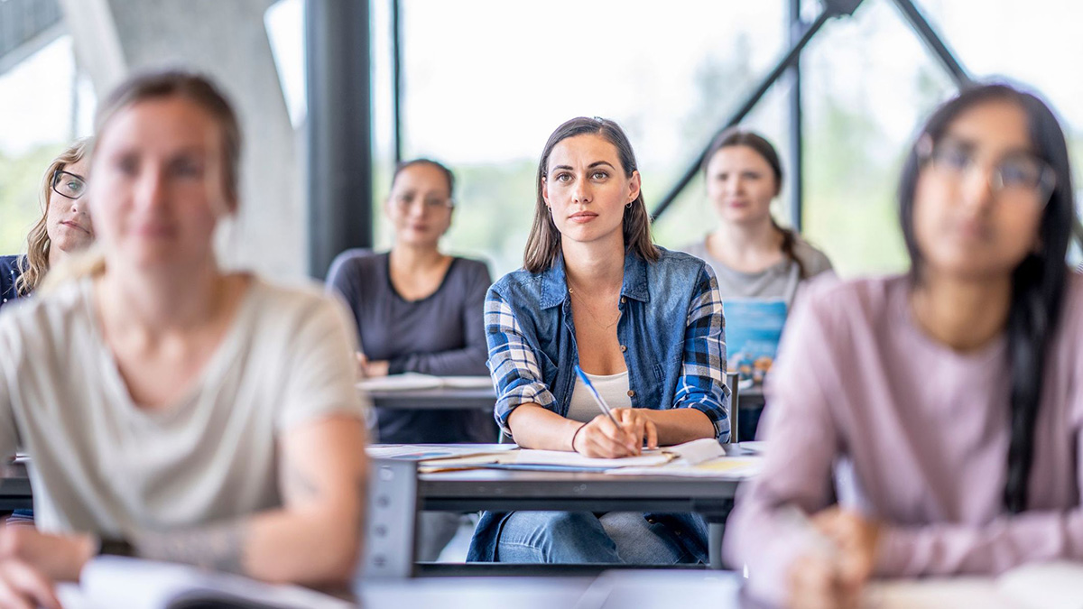 Gruppe von Studierenden sitzt in einem hellen Seminarraum; eine junge Frau in der Mitte macht sich Notizen und blickt aufmerksam nach vorne.